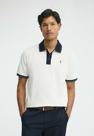 Homme portant un polo à manches courtes blanc avec col et bordure des manches bleu marine, pantalon bleu marine et ceinture marron, debout les mains dans les poches.