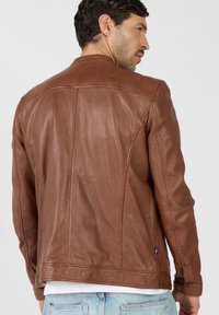 Ein Mann trägt eine taillierte braune Lederjacke, gesehen von hinten, kombiniert mit hellblauen Jeans und einem weißen Hemd darunter.
