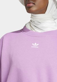 Fialová mikina s rebrovaným okrúhlym výstrihom a bielym logom Adidas na hrudi. Mäkkučký materiál s hladkým povrchom, preložený bielym šálom.