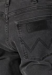 Posnetek iz bližine zadnjega žepa črnih jeans hlač z logotipom Wrangler in dekorativnim šivom na tkanini.
