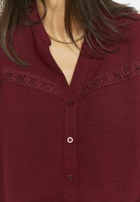 Bordo värvi pluus nööbitava esiküljega, millel on õrn pitsiline ääris rinna kohal ja lühikesed varrukad. Ühtlane tekstuur peene läikega.