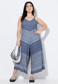 Overall a righe nei toni del blu e bianco, con scollo a V e gambe larghe. Presenta una tasca laterale ed è abbinato a una borsa silver e sandali con plateau.
