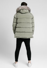 Giubbotto imbottito verde tenue con cuciture orizzontali, cappuccio foderato in pelliccia e pantaloni aderenti neri, completati da sneakers bianche.