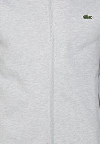 Sudadera gris con cremallera, de textura suave, con un logo de cocodrilo bordado en verde en el lado izquierdo del pecho y una cremallera central.