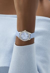 Relógio azul de silicone com caixa redonda prateada, detalhes em cristal, mostrador azul e marcadores com numerais romanos, usado num pulso com um casaco de malha azul claro.