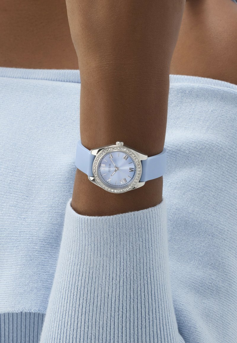 Relógio azul de silicone com caixa redonda prateada, detalhes em cristal, mostrador azul e marcadores com numerais romanos, usado num pulso com um casaco de malha azul claro.