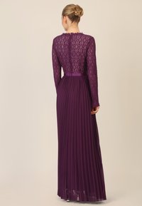 Pitkä violetti mekko, jossa on pitsinen yläosa, jossa on timanttikuvio ja pitkät hihat, sekä pleatattu hameosa ja vyötärö. 