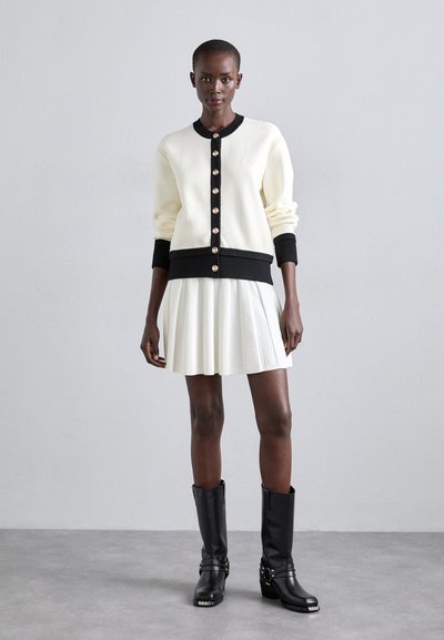 Cardigan crème avec des bordures noires, doté de boutons dorés et de poignets côtelés. Associé à une jupe plissée blanche et des bottes noires jusqu'aux genoux.
