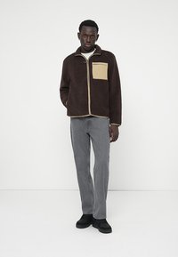 Mann steht in einer dunkelbraunen Fleecejacke mit beigem Taschen, grauen Jeans und schwarzen Schuhen vor einem weißen Hintergrund.