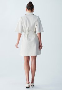 Heledad beež mantel dress lühikeste varrukatega, kirjakohaga kinni tõmbatud talje ja sirge lõikega, valmistatud tekstuursetest kangast. Seljas on valged kontsad.