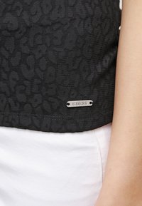 Zwarte tanktop van stof met een textuur van luipaardprint, inclusief een zilveren "GUESS" logo-label aan de zoom.
