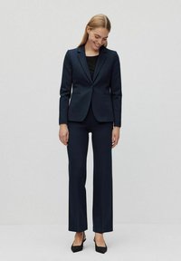 Marinblå kostym med figursydd kavaj och vida byxor, tillverkad av ett mjukt tyg. Kombineras med en svart topp och svarta högklackade skor.
