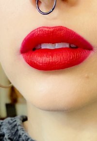 Red matte lipstick, smooth texture, defined shape with a slight gradient; septum ring visible. Natural skin tone background.