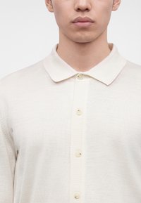 Jeune homme portant un polo en maille crème boutonné avec un col, montré du cou à la partie supérieure de la poitrine sur un fond uni.