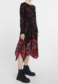Robe noire à manches longues avec motifs floraux rouges, ourlet asymétrique et détails superposés. Portée avec des bottines noires.