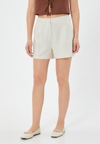 Shorts beiges taille haute avec une coupe ajustée, plis devant et texture lisse. Associés à des ballerines crèmes et un top court marron.