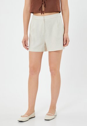 Beige højtaljede shorts med skræddersyet pasform, frontfolder og glat tekstur. Kombinationen med lyse creme ballerinasko og en brun cropped top.