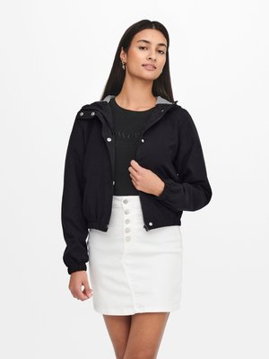 Mujer con cabello largo y oscuro que lleva una chaqueta negra con cremallera sobre una camiseta negra y una falda blanca con botones al frente, posando frente a un fondo blanco.