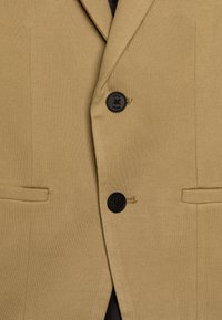 Barna blazer két fekete gombbal, strukturált hajtókával és sima, texturált anyaggal. Oldalsó zsebekkel és diszkrét logó részlettel rendelkezik.