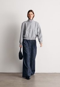 Grijze bomberjack met rits gecombineerd met losse, getailleerde marineblauwe broek, aangevuld met een kleine zwarte handtas en zwarte schoenen.