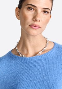 Blauw wollen trui met een geribbelde textuur en ronde halslijn, gecombineerd met een dikke zilveren ketting met gestructureerde schakel.