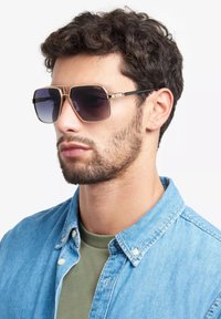 Occhiali da sole quadrati con lenti scure sfumate, dettagli in metallo dorato e montatura nera. Il modello indossa una camicia in denim sopra una maglietta verde.