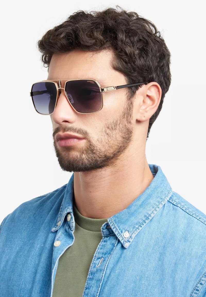 Occhiali da sole quadrati con lenti scure sfumate, dettagli in metallo dorato e montatura nera. Il modello indossa una camicia in denim sopra una maglietta verde.