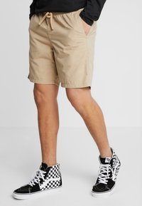Beige Shorts mit elastischem Bund und seitlichen Taschen, kombiniert mit schwarz-weißen karierten High-Top-Sneakern. Strukturiertes Material mit einer lockeren Passform.