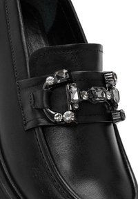 Zwarte leren loafers met een decoratieve gesp versierd met verschillende zwarte en heldere edelstenen, die een gladde textuur en een slank ontwerp tonen.