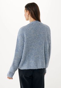 Kobieta z długimi brązowymi włosami, nosząca srebrne kolczyki w kształcie kół, niebieski sweter o teksturowanym wzorze i czarne spodnie, zwrócona plecami na tle białym.