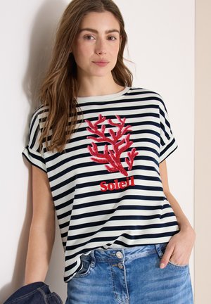 Frau trägt ein schwarz-weiß gestreiftes Shirt mit rotem Korallendesign und dem Schriftzug "Soleil", kombiniert mit blauer Jeans, steht an einer Wand.