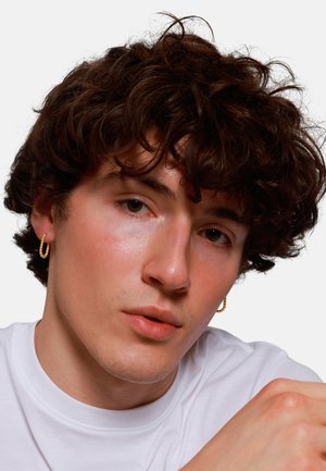 Gouden creolen met een textuurdesign, gedragen door een persoon met krullend donker haar, in een witte T-shirt, met een neutrale uitstraling.