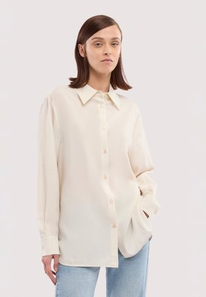 Kobieta o prostych brązowych włosach, ubrana w luźną, kremową koszulę na guziki i jasnoniebieskie dżinsy, stojąca na tle jednolitego tła.