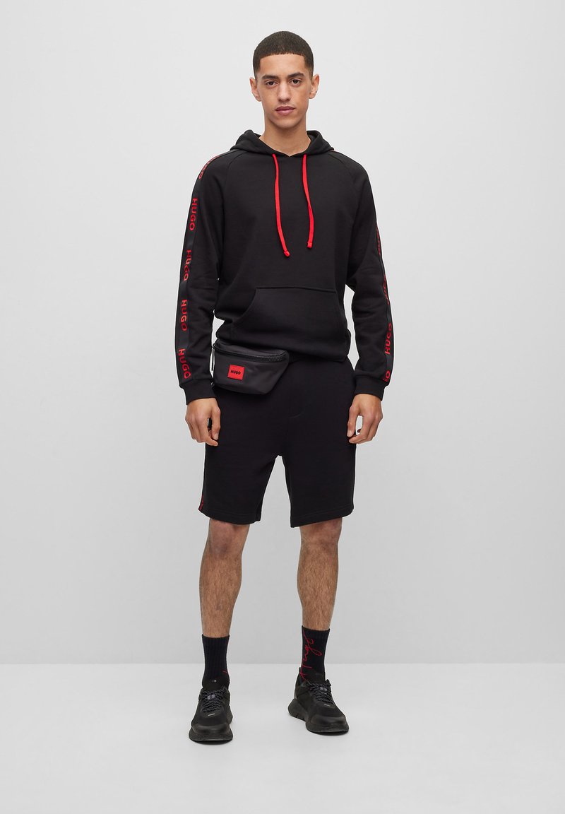 Svart hettegenser med røde detaljer og snorer, kombinert med svarte shorts og svarte joggesko. Svart høypotetveske brukt over hoftene.