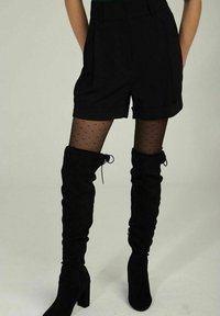 Shorts noirs ajustés avec plis, associés à des collants noirs transparents à pois et des bottes noires en daim montant au-dessus des genoux avec talons carrés.