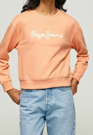 Persona con sudadera color melocotón con texto "Pepe Jeans" y vaqueros de mezclilla azul claro, con las manos apoyadas en las caderas frente a un fondo liso.