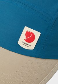 Niebieska czapka z materiału z kremowym daszkiem. Posiada naszywkę z logo czerwonego lisa oraz etykietę "FJÄLLRÄVEN", wzbogaconą widocznymi detalami przeszycia.
