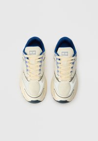 Adidași cu un corp crem, accente albastre și detalii din plasă. Au șireturi beje și o talpă curbată pentru un plus de confort. Logo-ul este afisat proeminent.