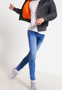 Chaqueta bomber negra con forro naranja, camiseta blanca y jeans ajustados azules. El modelo lleva zapatillas grises y posa en una postura dinámica.