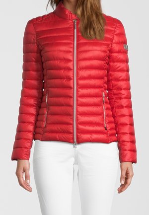 Femme portant une veste matelassée rouge ajustée avec une fermeture éclair devant et des poches latérales zippées, associée à un pantalon blanc, debout devant un fond uni.