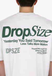 T-shirt en coton blanc avec du texte vert. Le dos affiche "DropSize" et d'autres phrases dans des tailles et styles de police variés. Le texte inclut des détails sur la marque.