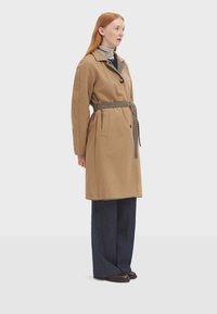 Trench coat beige con cintura ceñida, que presenta un cinturón y un cuello a cuadros. Llevado sobre un suéter gris de cuello alto y pantalones anchos oscuros.