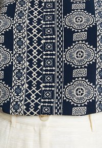 Námořnická košile s bílými geometrickými a květinovými vzory, vyrobená z texturované látky, spárovaná s světlými kalhotami.