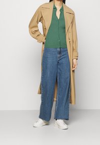 En kvinna bär en grön ribbad kofta, ljusblå vida jeans och en beige trench coat, stående mot en enfärgad bakgrund.