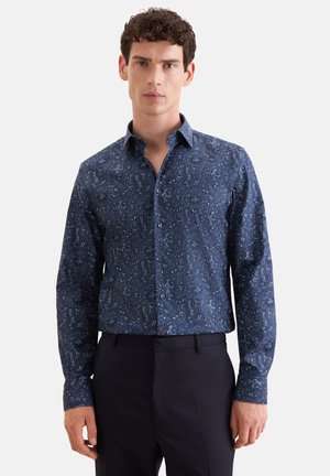 Homme aux cheveux bouclés portant une chemise bleu foncé à manches longues avec un petit motif floral et un pantalon noir, debout face à nous.