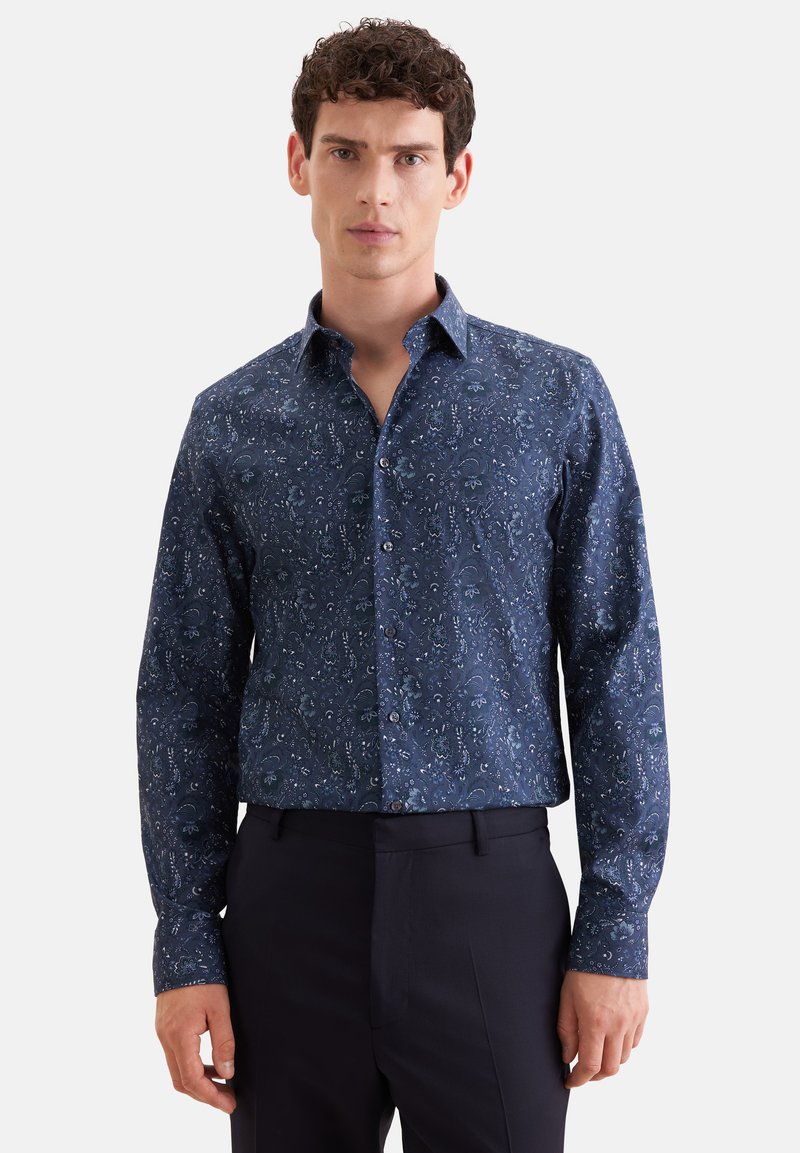 Homme aux cheveux bouclés portant une chemise bleu foncé à manches longues avec un petit motif floral et un pantalon noir, debout face à nous.