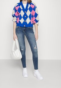 Cárdigan de punto con patrón de rombos en azul, rosa y beige, con escote en V y botones dorados, combinado con vaqueros azules desgastados y zapatillas blancas.