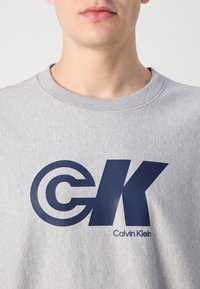 Šedá bavlněná mikina s výrazně vytištěným tmavě modrým logem "CK" a textem "Calvin Klein". Design s kulatým výstřihem, měkká textura.