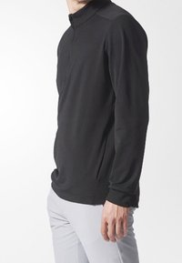 Hombre vestido con un pulóver negro de manga larga con cremallera en el cuello y pantalones gris claro, de pie contra un fondo blanco.