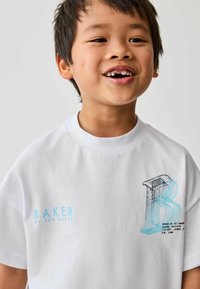 Bílý bavlněný tričko s velkým modrým obrysovým písmenem 'B' a světle modrým textem "BAKER BY TED BAKER" na přední straně. Kulatý výstřih, krátké rukávy.